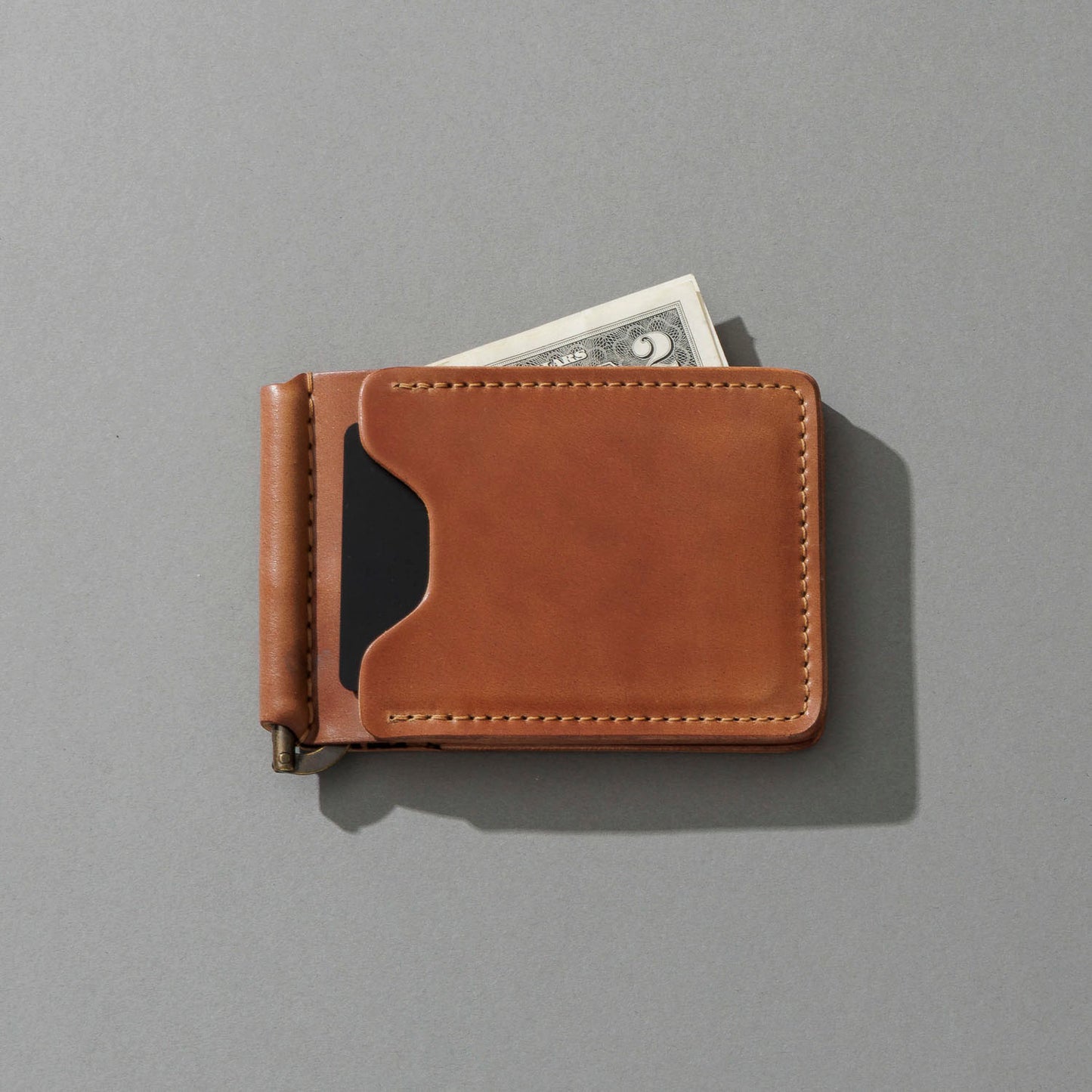 A tan leather money clip wallet with a black card slot, shown closed with a dollar bill partially tucked into one of its card slots, on a solid gray background.
