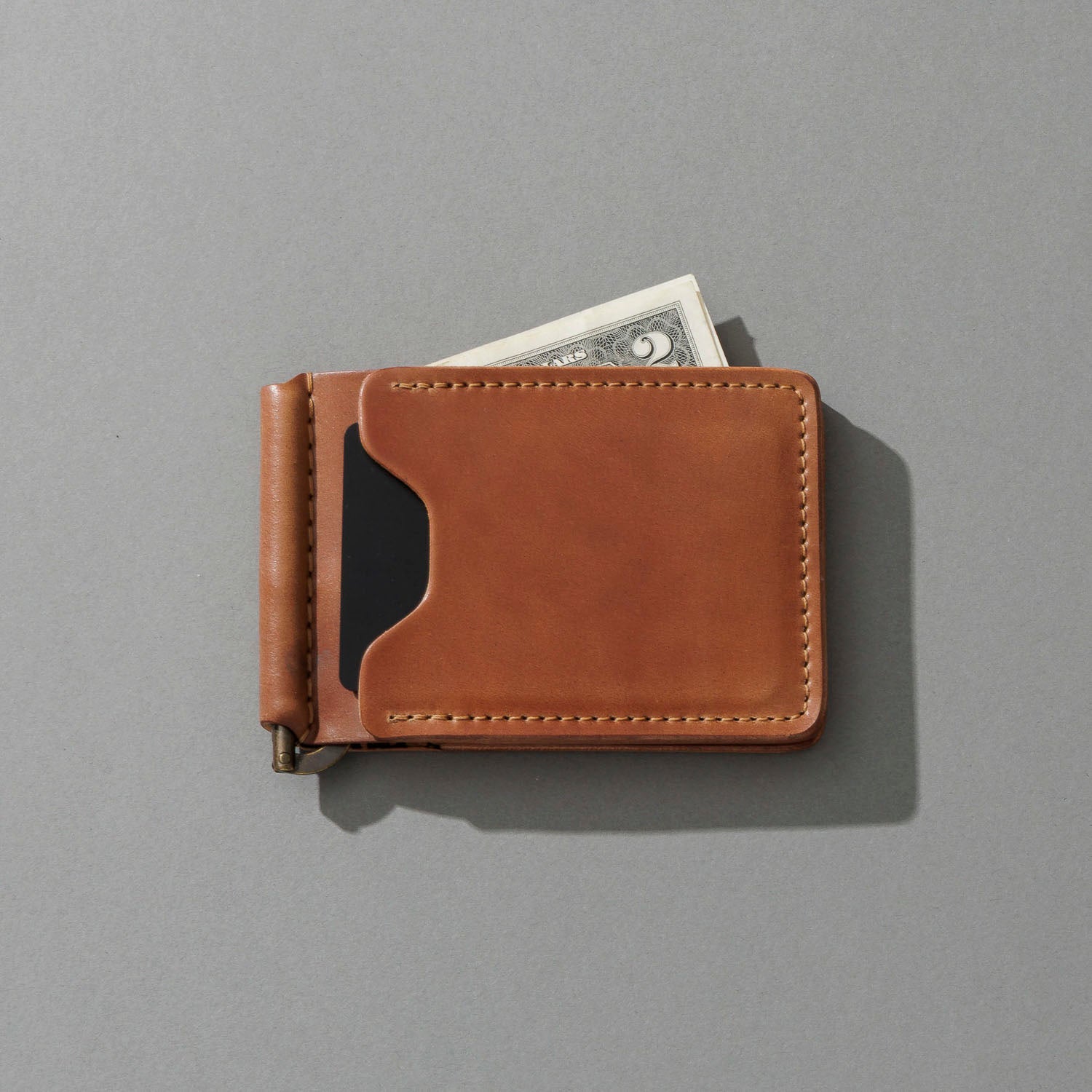 A tan leather money clip wallet with a black card slot, shown closed with a dollar bill partially tucked into one of its card slots, on a solid gray background.