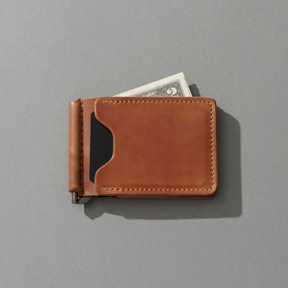 A tan leather money clip wallet with a black card slot, shown closed with a dollar bill partially tucked into one of its card slots, on a solid gray background.