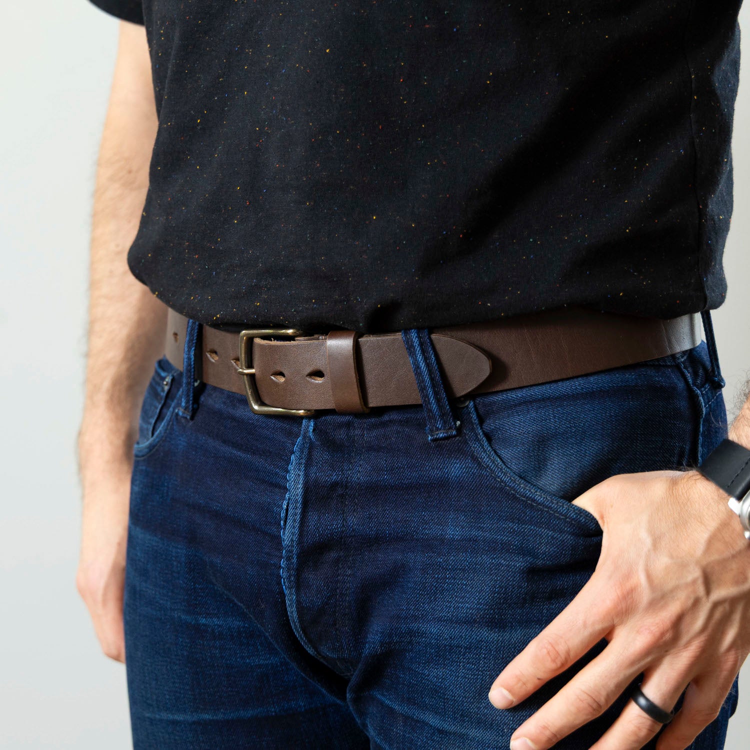 Person wearing a black shirt, blue jeans, and a brown belt on a plain background