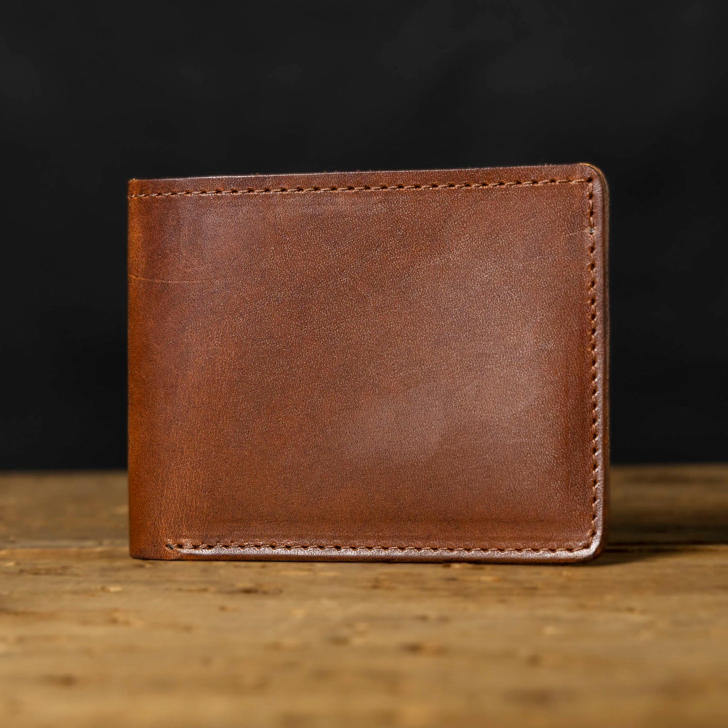 A brown bifold leather wallet with visible stitching and a logo embossed on the bottom right. The wallet is displayed in an open and closed position on a wooden surface.