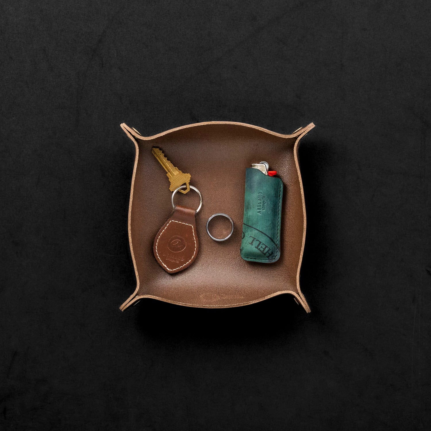 Brown leather tray with keychain, ring, and lighter on a black background